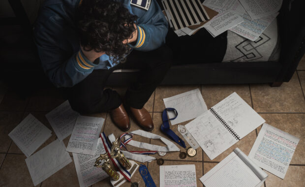 Photograph of young person sitting at the edge of a bed with a view of school papers, awards, and other items on the ground. The person looks distressed, and they're facing down away from the camera.
